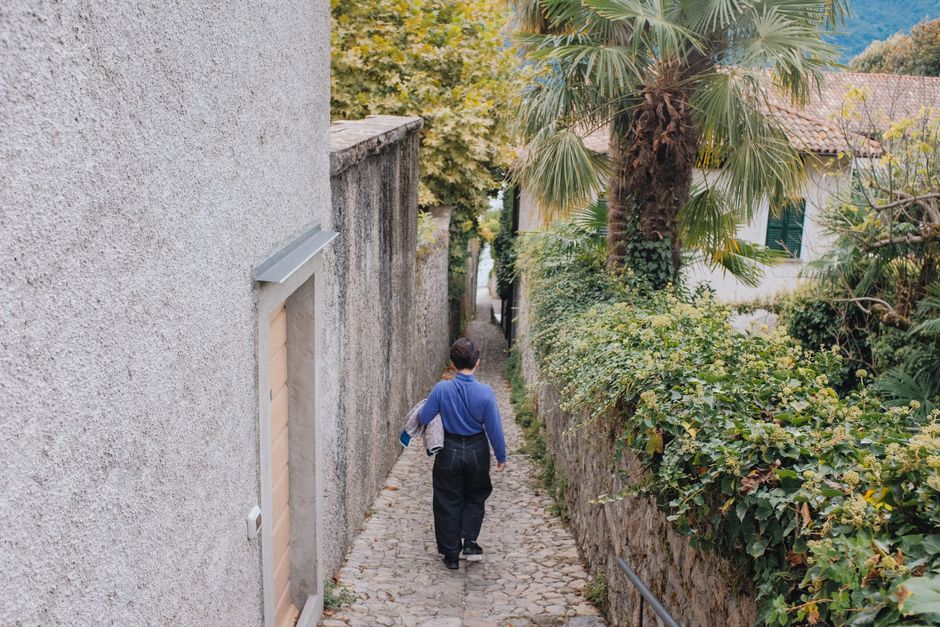 A person walks down a narrow cobblestone alley surrounded by lush greenery in a picturesque European setting.